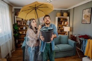 A woman holding an umbrella and a man holding a bucket indoors for a roof leak emergency by NorCal Roofing & Gutters