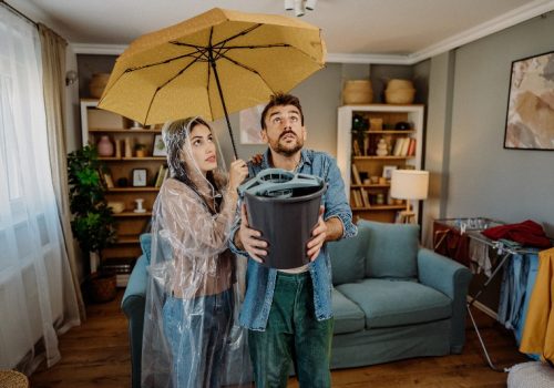 A woman holding an umbrella and a man holding a bucket indoors for a roof leak emergency by NorCal Roofing & Gutters